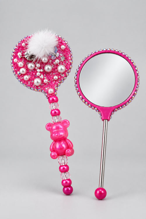 A pink decorative hand mirror with a beaded handle and pom-pom top, displayed next to a matching pink rhinestone-lined case on a marble surface.